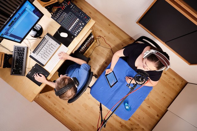 Blick ins audiacc-Studio aus der Vogelperspektive. Eine Studio-Sprecherin sitzt an einem quadratischen Tisch mit einer blauen Tischdecke. Sie trägt einen Studio-Kopfhörer, liest Text von einem Tablet ab und spricht in ein Studio-Mikrofon.