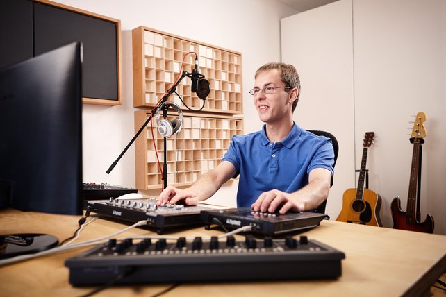 Florian Schmitz sitzt am Studio-Tisch und schaut in Richtung des Computer-Monitors. Im Vordergrund ist die Studio-Technik zu sehen. An der Wand sind Akustik-Module montiert und im Hintergrund stehen ein E-Bass und eine Akustik-Gitarre.